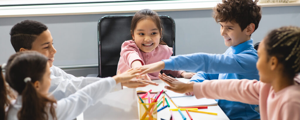 Education, Elementary School, Children, Break and People Concept. Diverse group of smiling school kids putting hands on top together sitting at table in classroom, young friends making team