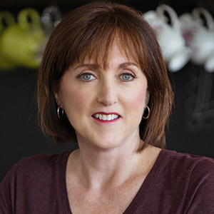 “Portrait of a middle-aged woman with short auburn hair, light skin, and blue eyes, smiling softly at the camera. She is wearing hoop earrings and a dark V-neck top. The background is softly blurred with teacups displayed behind her.
