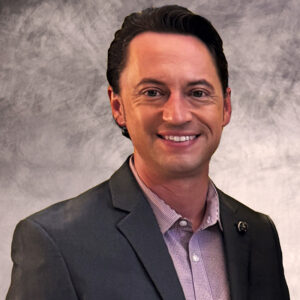 Professional headshot of a smiling man wearing a dark blazer and collared shirt against a gray textured studio background.